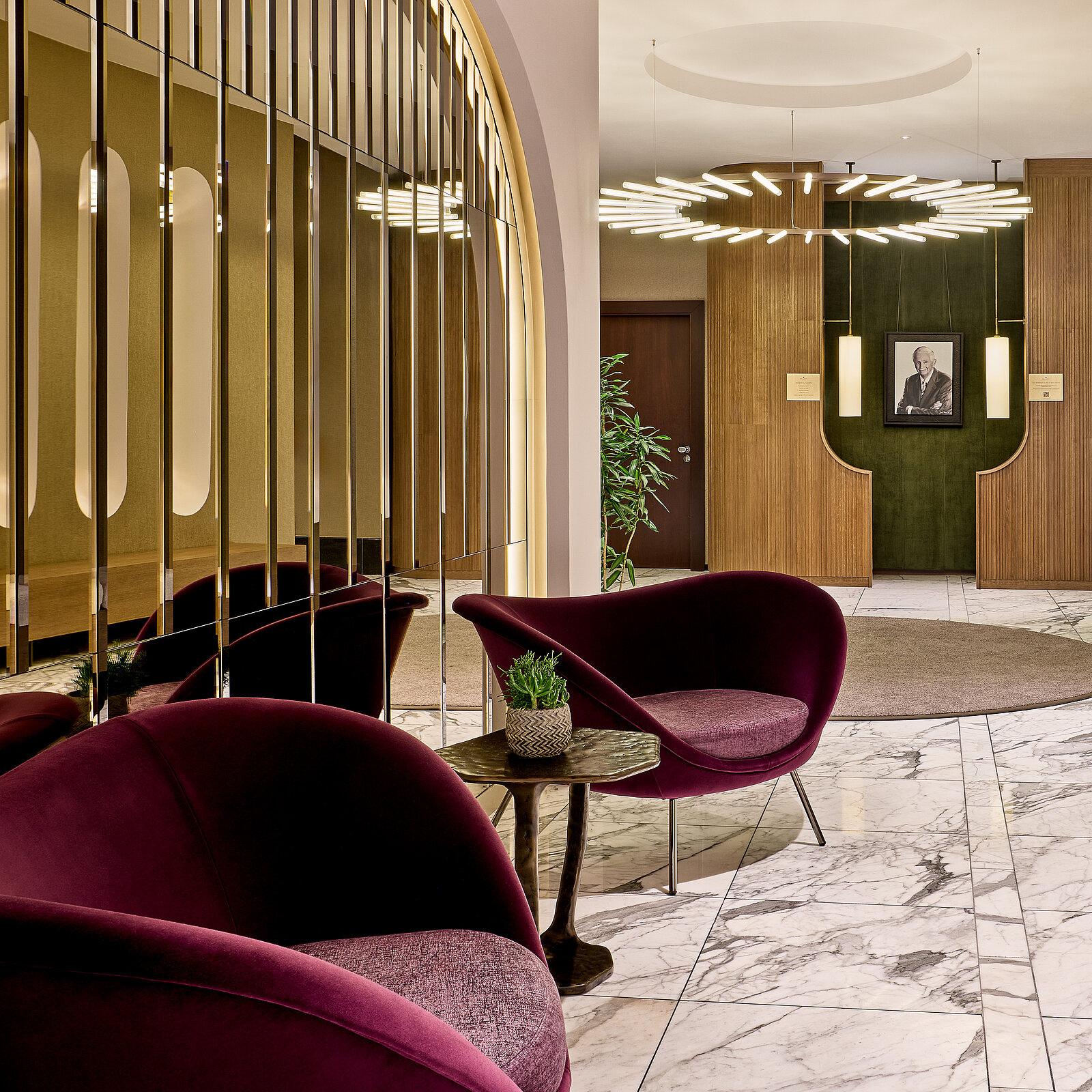 In einer eleganten Lobby mit Marmorboden stehen zwei bordeauxrote Sessel und ein kleiner Tisch. An der Wand befindet sich ein Gemälde. Eine große, moderne Deckenleuchte hängt in der Mitte des Raumes.