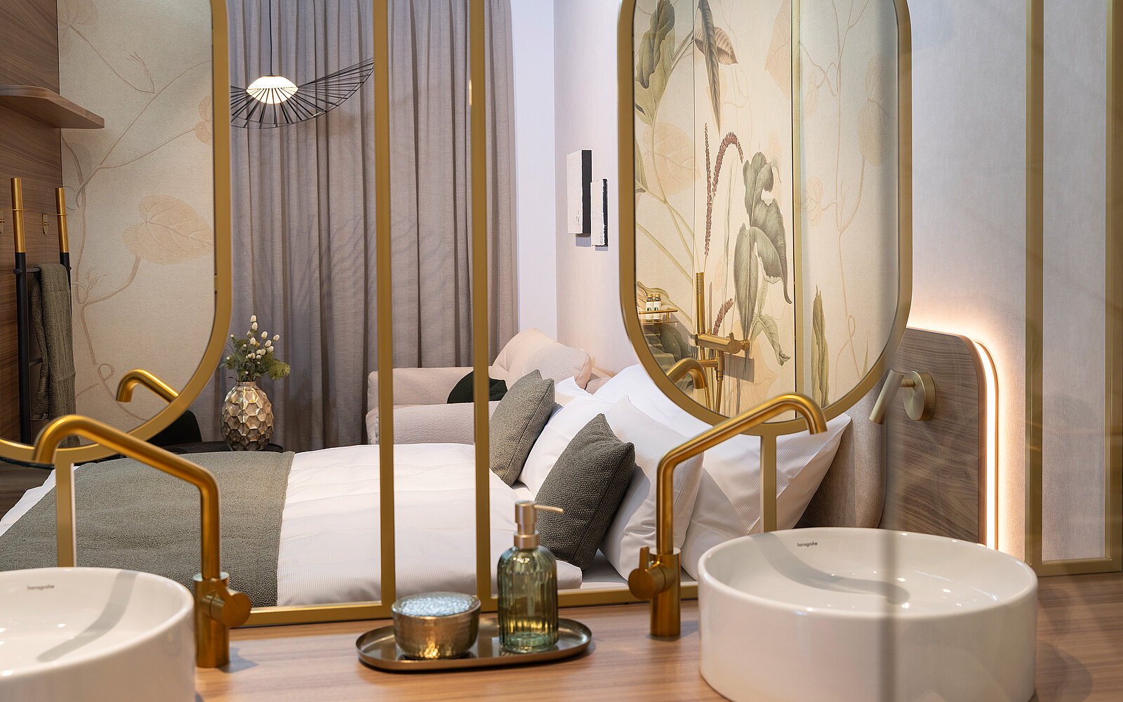 A modern bedroom is visible through a decorative frame. The room features a large bed with white bedding, soft pillows, and elegant wall decor of leaves. In the foreground, two white sinks sit on a wooden surface, accompanied by a small tray holding a bottle and a plant.