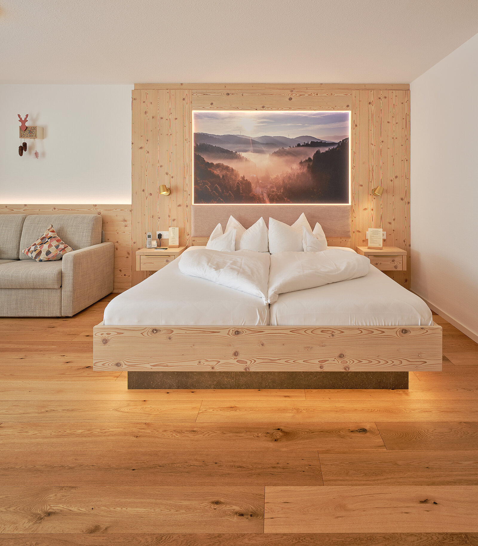 Ein gemütliches Schlafzimmer mit Holzboden und Holzwand. Ein großes Bild von Bergen bei Sonnenaufgang hängt über dem Doppelbett. Neben dem Bett stehen zwei Nachttische mit Lampen. Links davon befindet sich ein graues Sofa. Der Raum ist stilvoll eingerichtet.