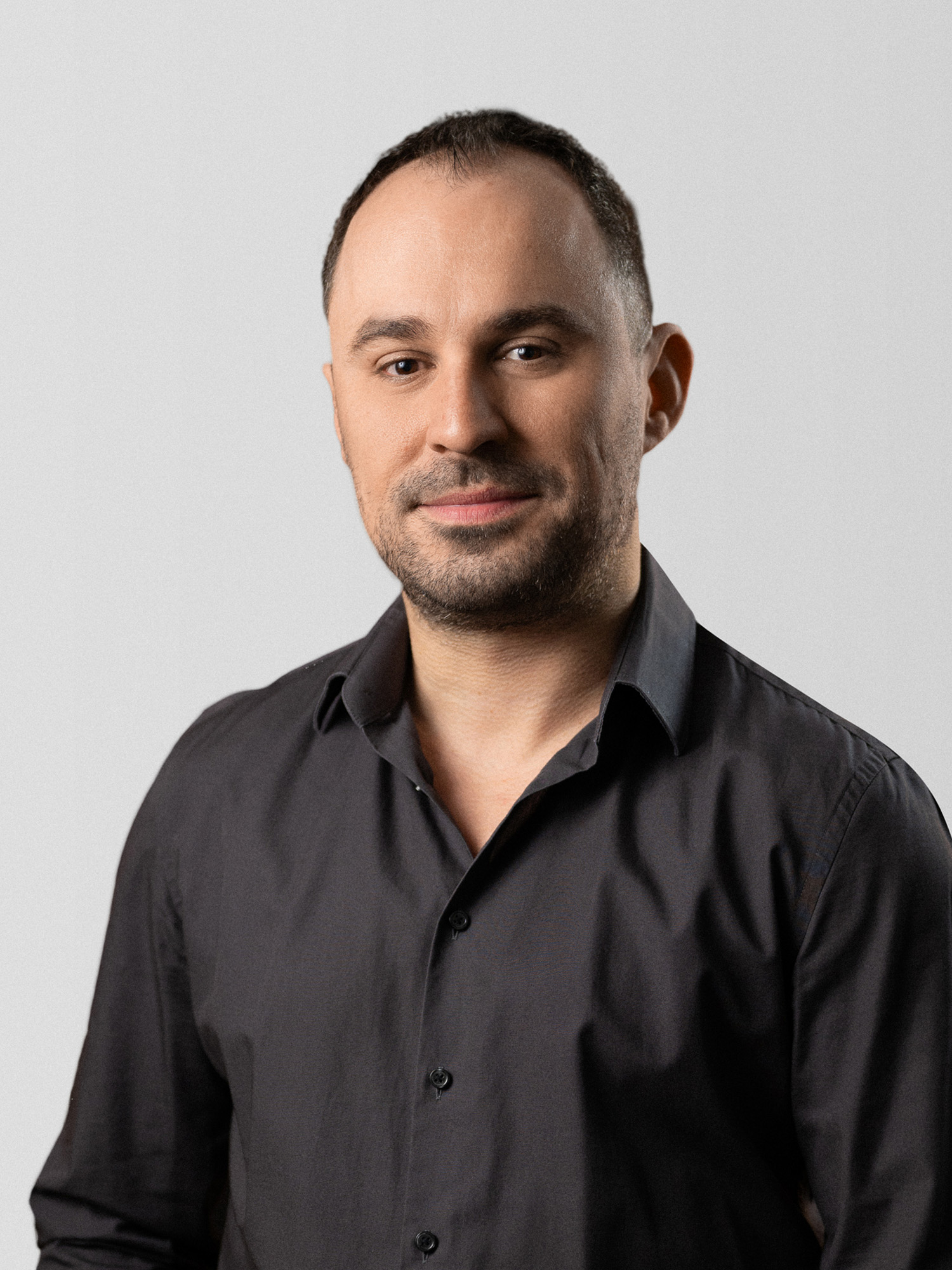 Ein Mann mit kurzen, dunklen Haaren steht vor einem hellen Hintergrund. Er trägt ein schwarzes Hemd und lächelt leicht. Sein Gesichtsausdruck ist freundlich und selbstbewusst. Die Aufnahme zeigt ihn von der Brust aufwärts.