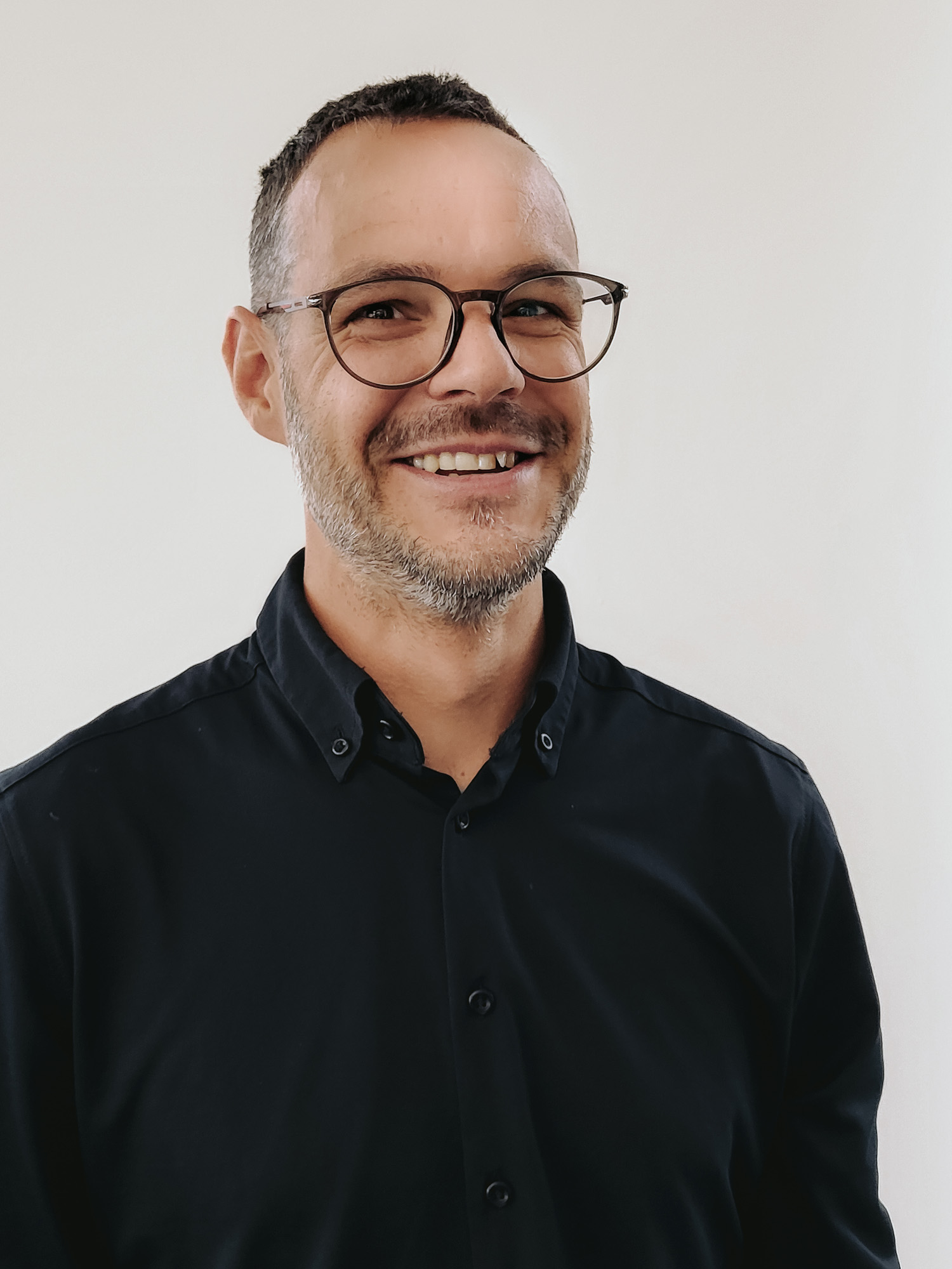 Ein lächelnder Mann mit Brille steht vor einem hellen Hintergrund. Er trägt ein schwarzes Hemd und hat kurzes, gepflegtes Haar. Der Ausdruck seines Gesichts vermittelt Freundlichkeit und Selbstbewusstsein.