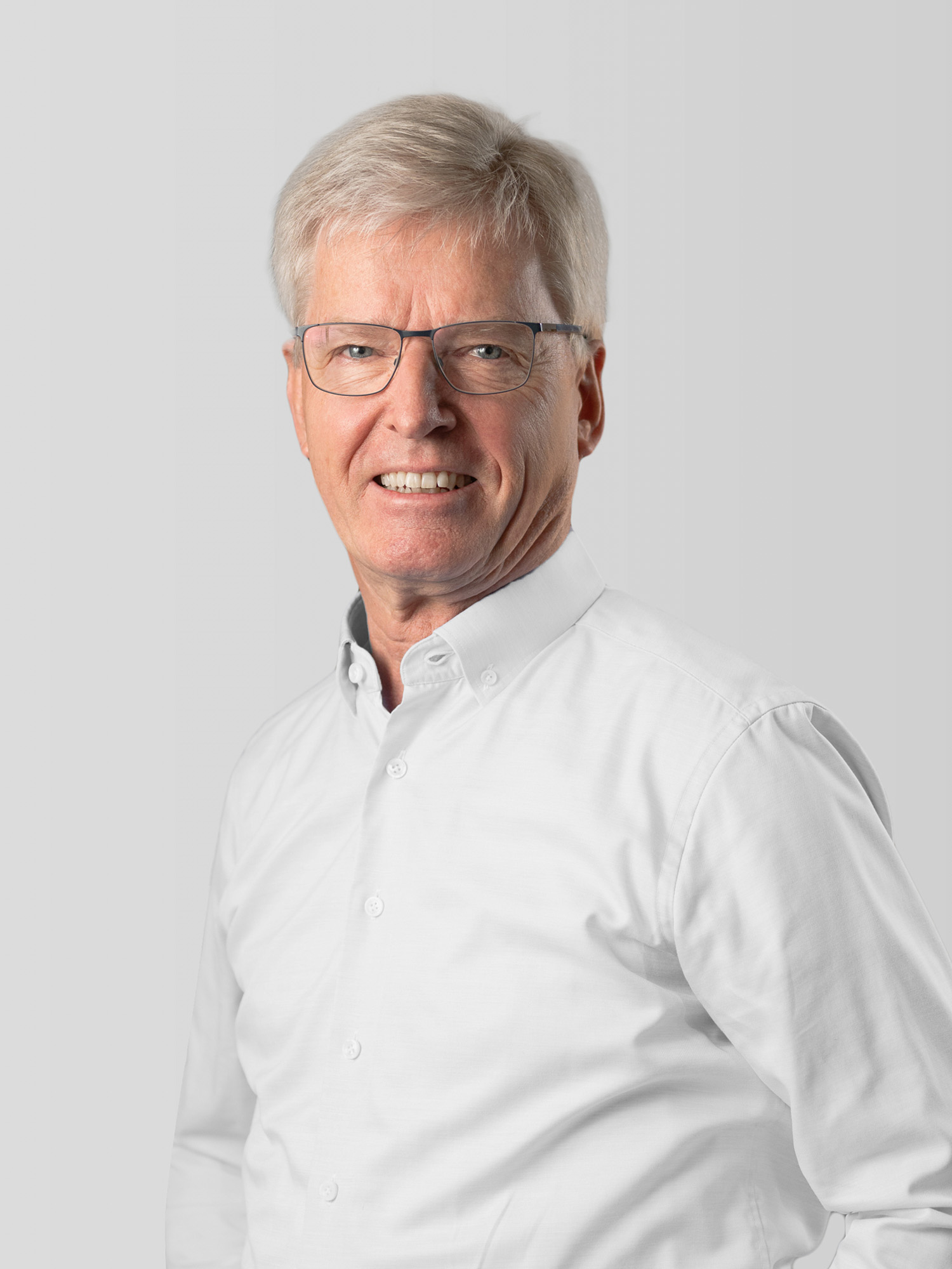 Das Bild zeigt einen älteren Mann mit kurzen, grauen Haaren, der in die Kamera lächelt. Er trägt eine weiße Hemdbluse und eine modische Brille. Der Hintergrund ist neutral und hell, was den Fokus auf sein freundliches Gesicht legt.