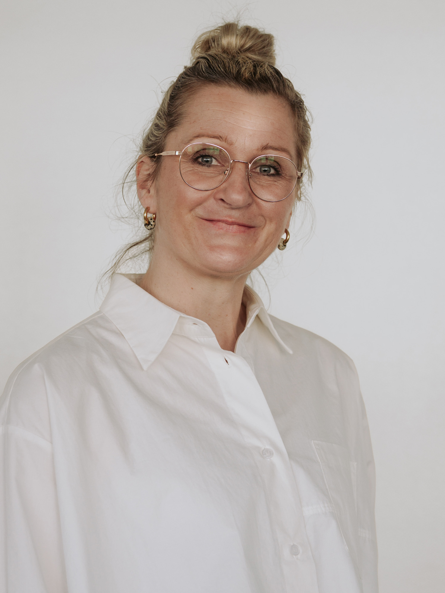 Ein Porträt einer lächelnden Frau mit blonden Haaren, die zu einem Dutt gebunden sind. Sie trägt eine weiße Bluse und eine goldene Brille. Der Hintergrund ist neutral. Die Frau hat große Ohrringe und strahlt eine freundliche Ausstrahlung aus.