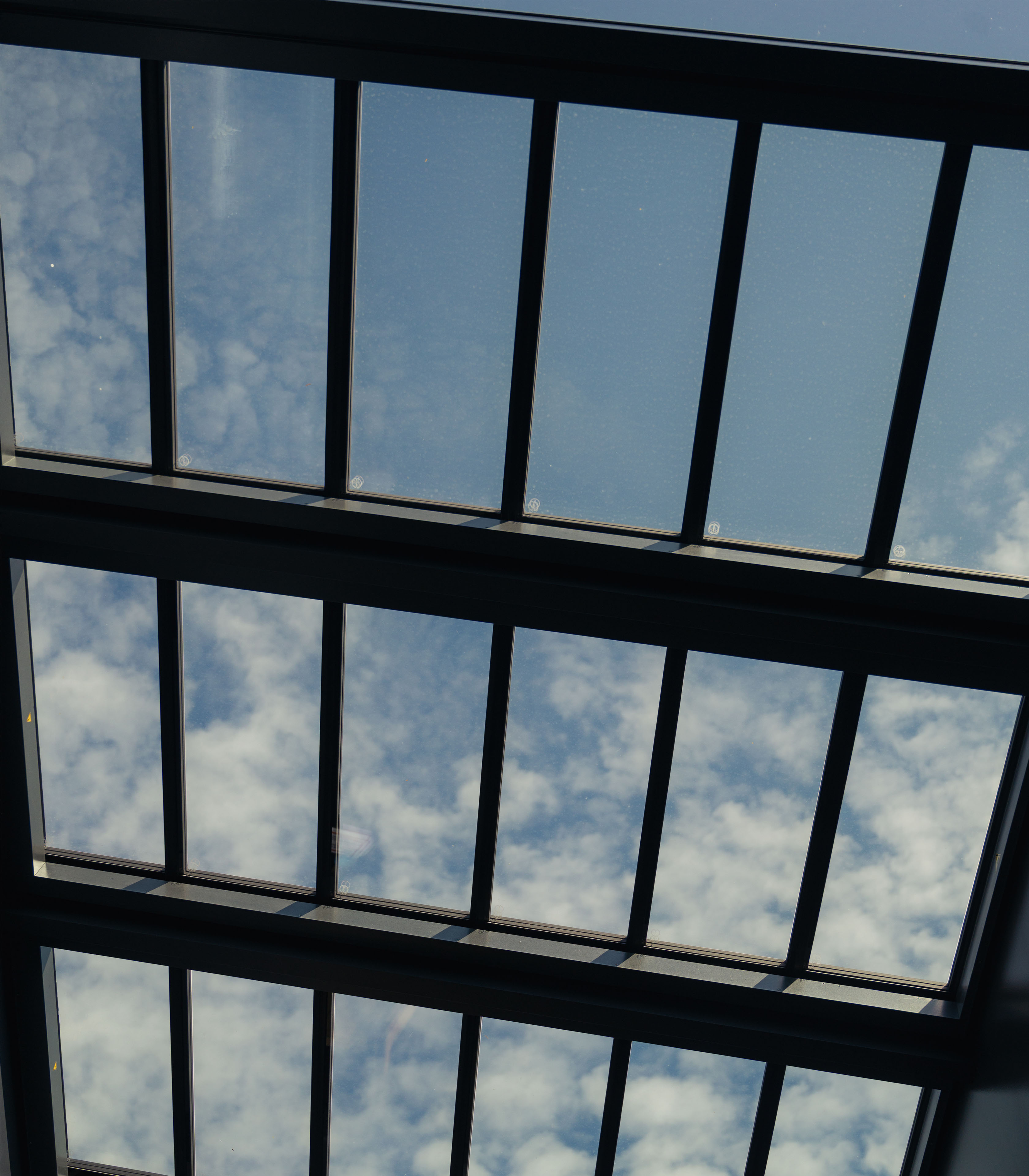 Eine Ansicht nach oben zu einem Oberlicht mit mehreren Glasfeldern. Die Scheiben sind schwarz gerahmt und geben den Blick auf den blauen Himmel frei, durch den weiche, weiße Wolken hindurchscheinen. Das Licht ist hell und schafft eine luftige, offene Atmosphäre.