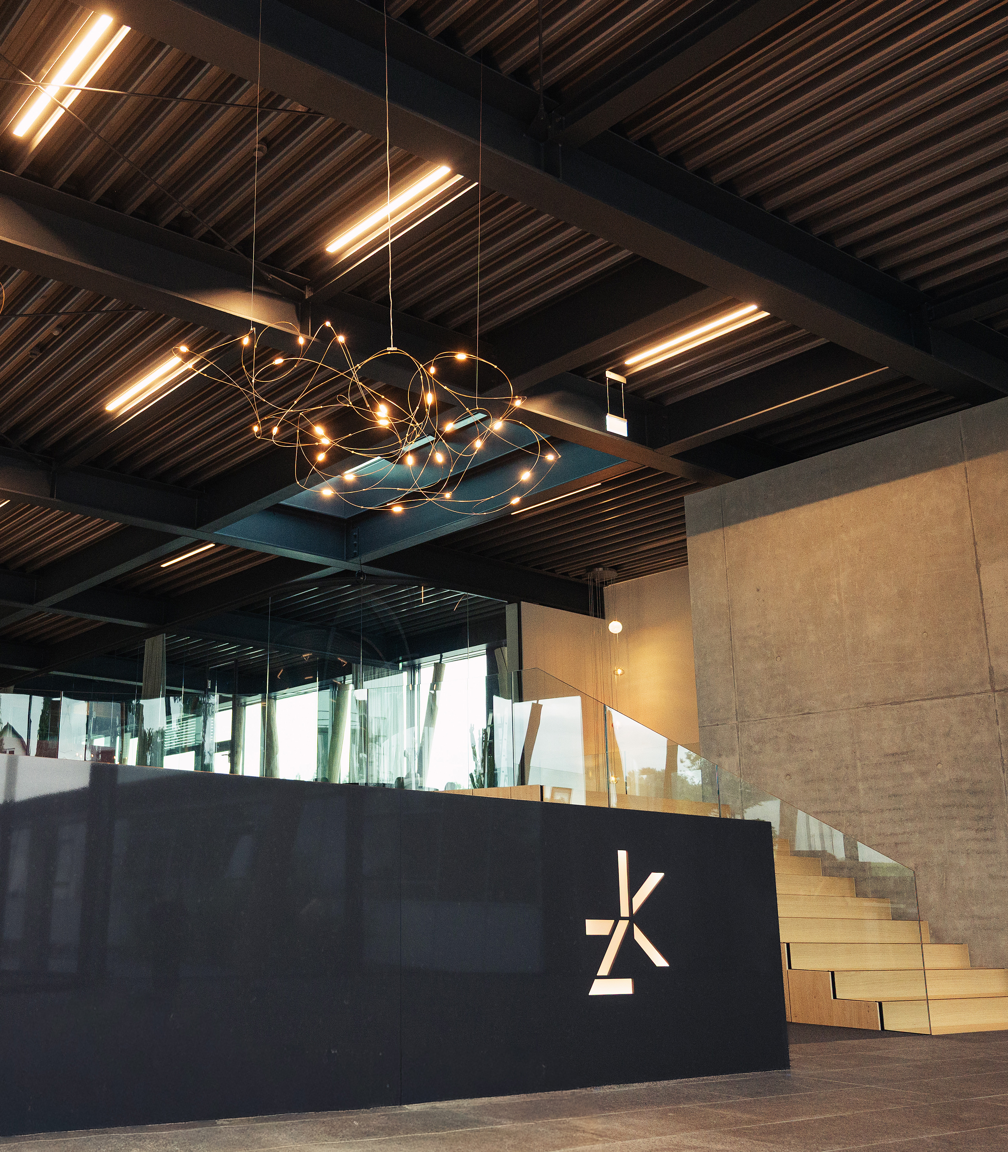 Das Bild zeigt die Empfangsbereiche eines modernen Gebäudes. Im Vordergrund steht ein dunkler Empfangstresen mit einem markanten Logo, dahinter ist eine lichtdurchflutete Glaswand sichtbar. Über dem Tresen hängt eine künstlerische Leuchte aus filigranen Linien.