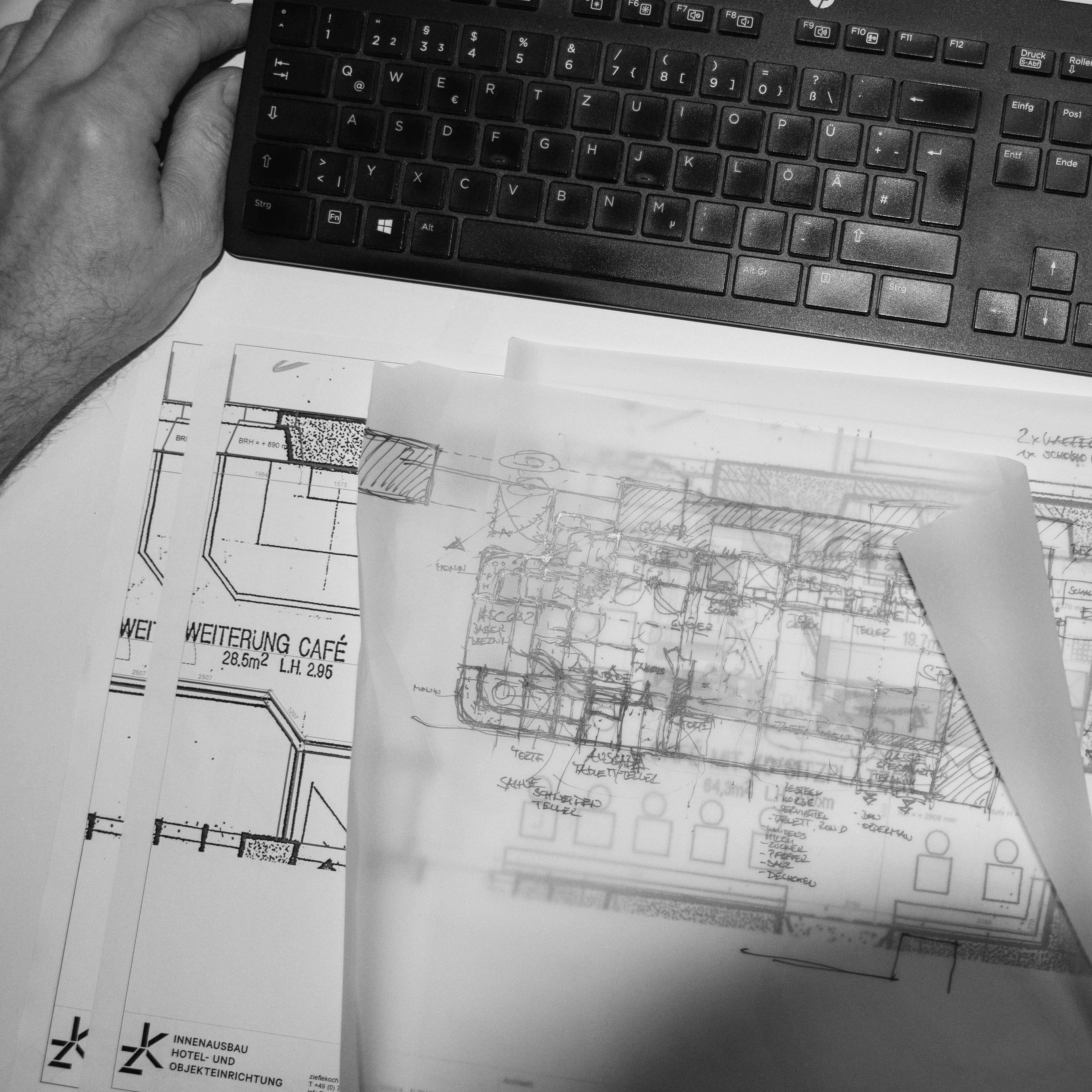 Ein Schreibtisch mit Plänen eines Café-Entwurfs, darüber eine Hand auf einer Tastatur. Architektonische Zeichnungen und Details des Innenausbaus sind sichtbar, die auf Erweiterungen hinweisen. Die Pläne sind beschriftet und teilweise überlappen sie sich.