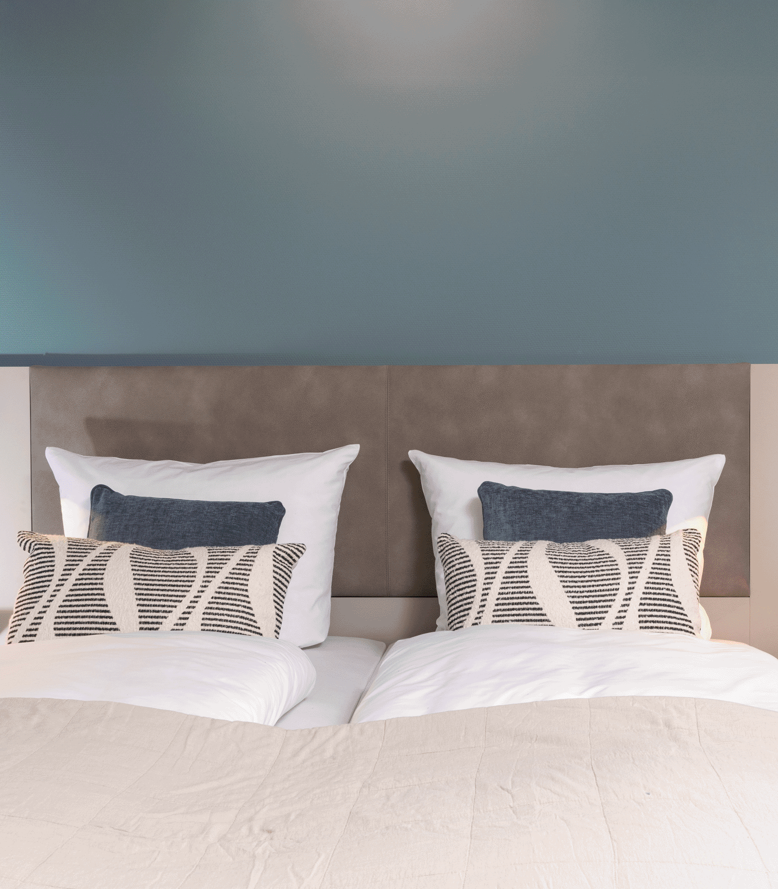 Ein gemütliches Doppelbett mit weißen Bettdecken und zwei Kissen. Die Kissen sind in verschiedenen Farben und Mustern gehalten. Im Hintergrund ist eine sanfte, blaue Wand zu sehen.