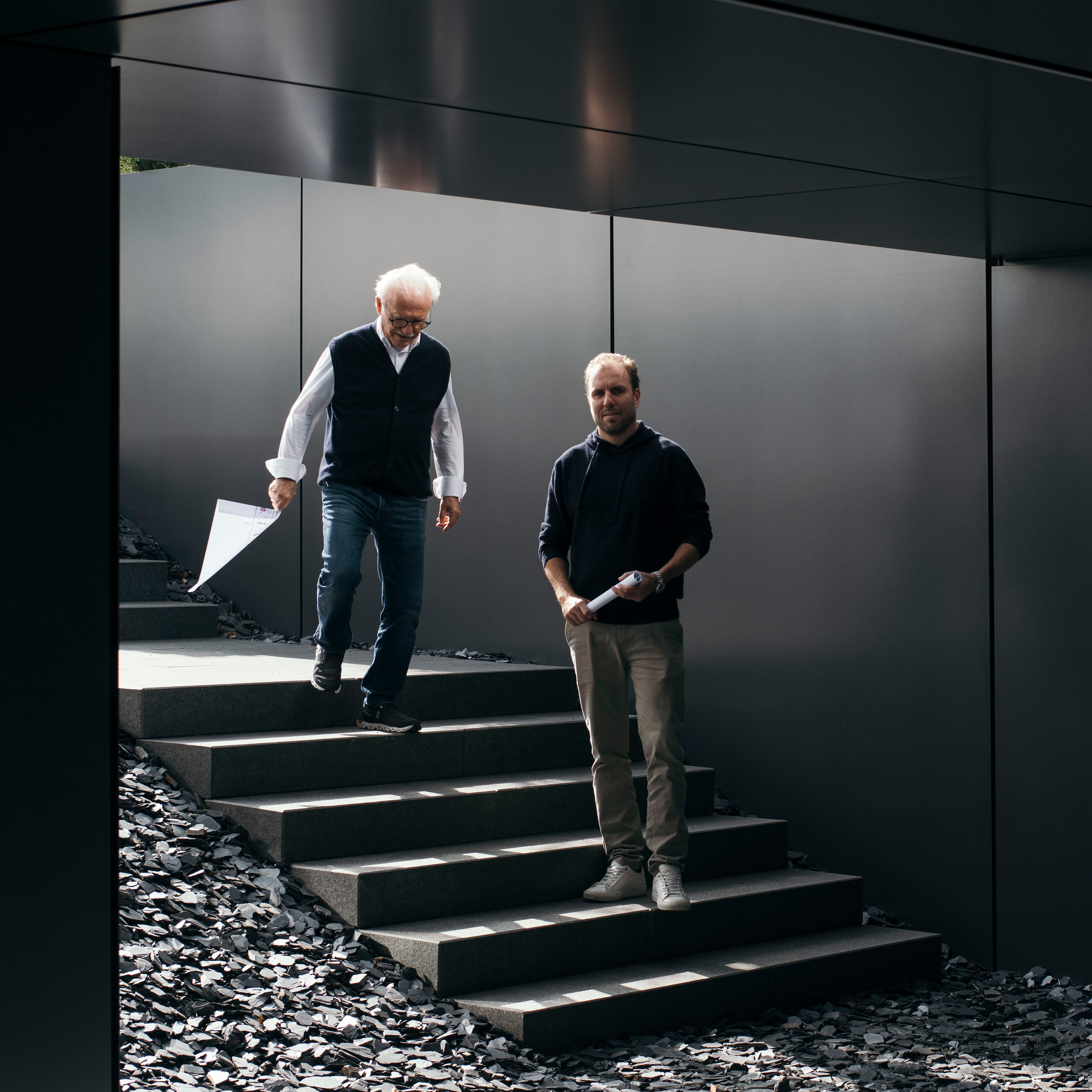 Two men are walking down a set of steps surrounded by dark stones. One man, with white hair, holds a piece of paper and wears a vest, while the other, with shorter hair, holds a phone and wears a dark sweater. They appear engaged in conversation.