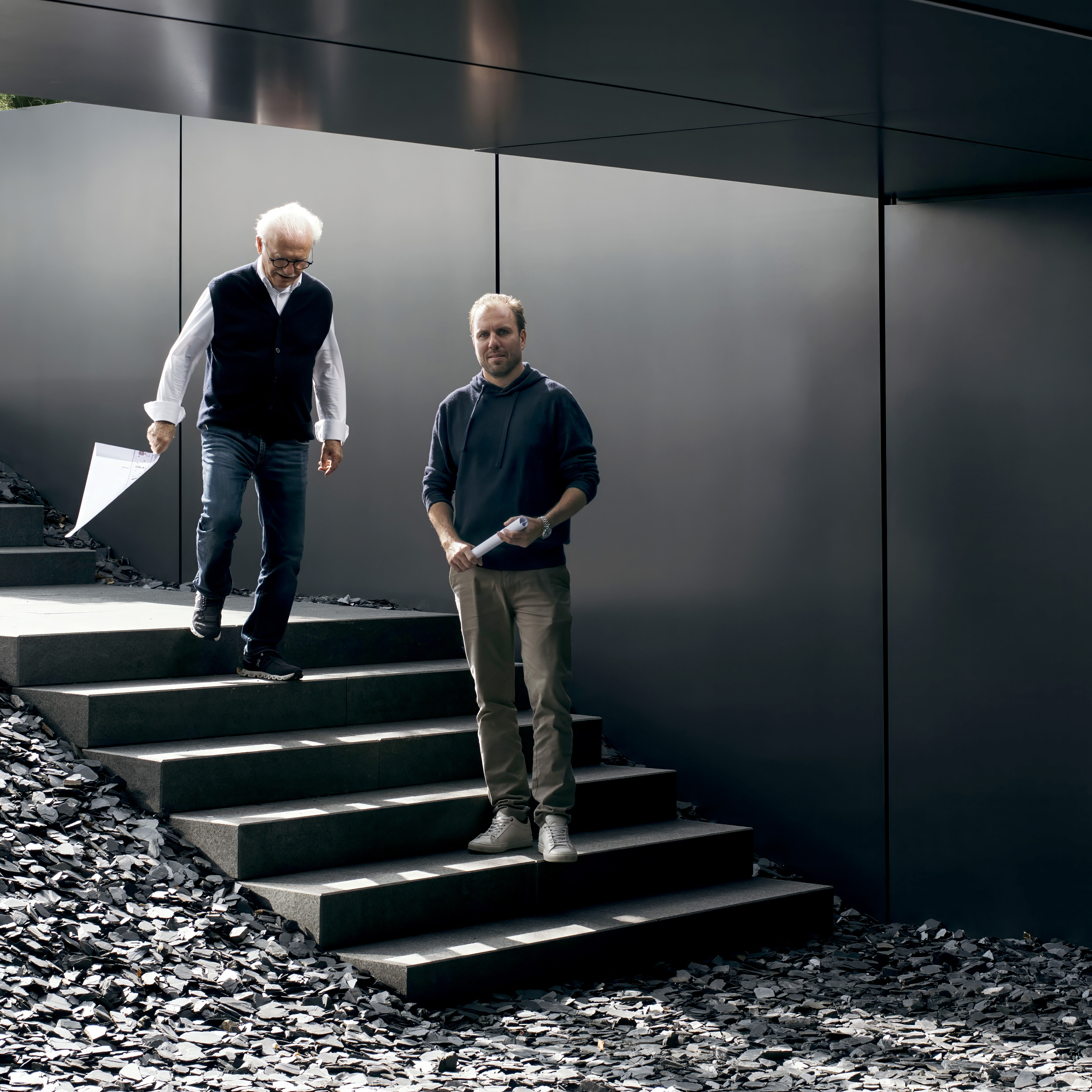 Two men are standing on a staircase made of large stone steps, surrounded by dark gravel. One man, older with white hair, is holding a paper, while the younger man stands beside him, looking at a watch. They appear to be engaged in conversation.