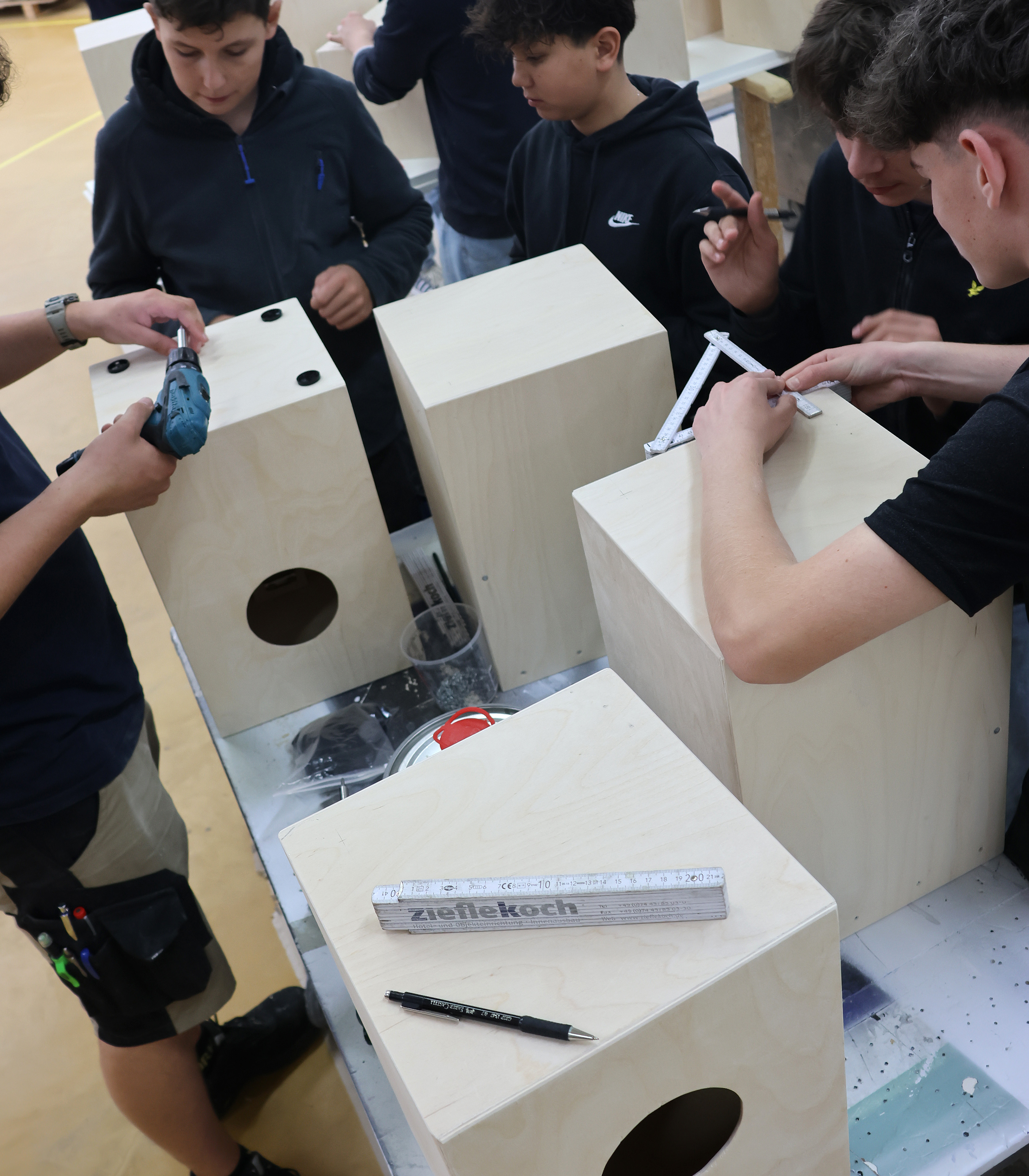 Eine Gruppe von Jugendlichen arbeitet an Holzkisten. Einige nutzen Werkzeuge, während andere Maße nehmen. Die Kisten haben runde Aussparungen und sind auf einem Tisch angeordnet. Es herrscht eine kreative, handwerkliche Atmosphäre.