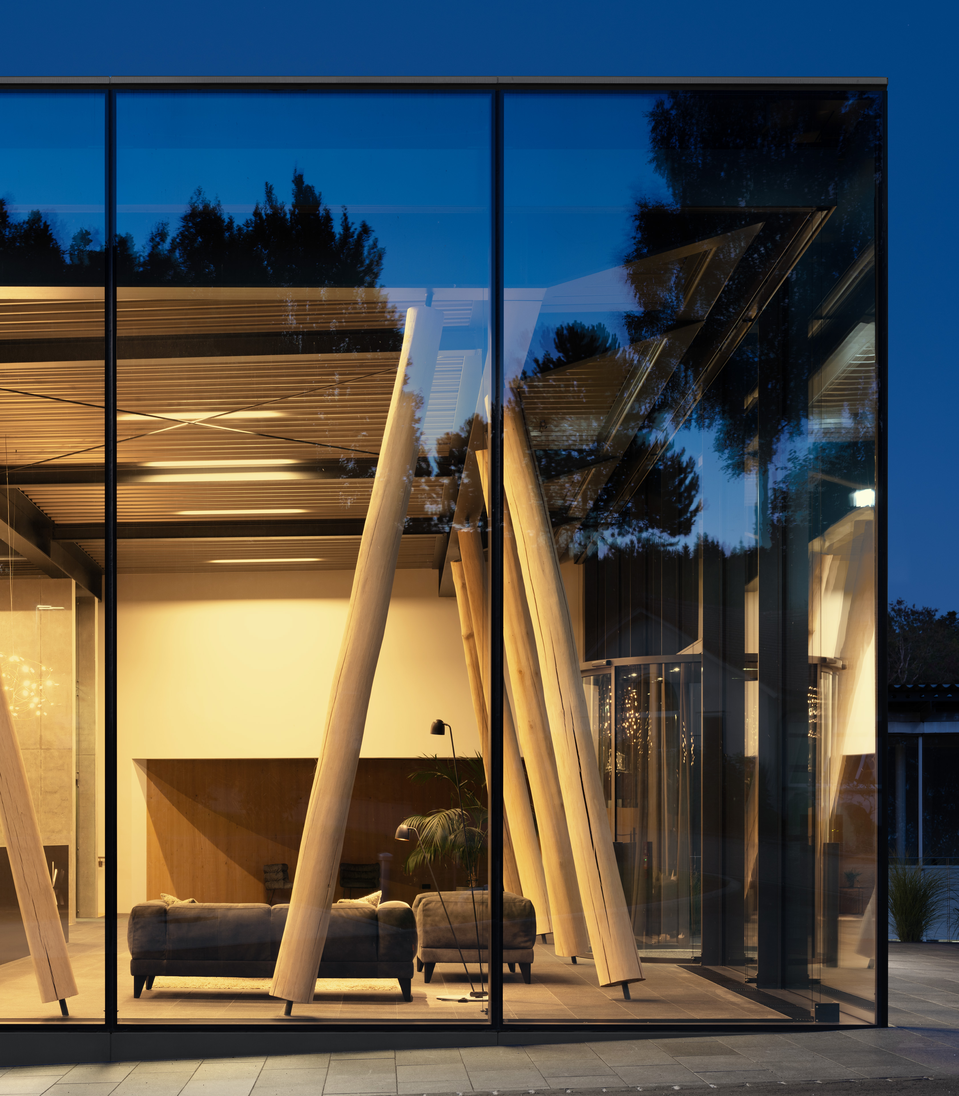 Eine moderne Konstruktion mit Glaswänden verfügt über freiliegende Holzbalken und eine gemütliche Inneneinrichtung mit einem Sofa und einer Pflanze. Die Szene wird in der Abenddämmerung beleuchtet, wodurch das Wechselspiel von Licht und Schatten innerhalb des Raums hervorgehoben wird.