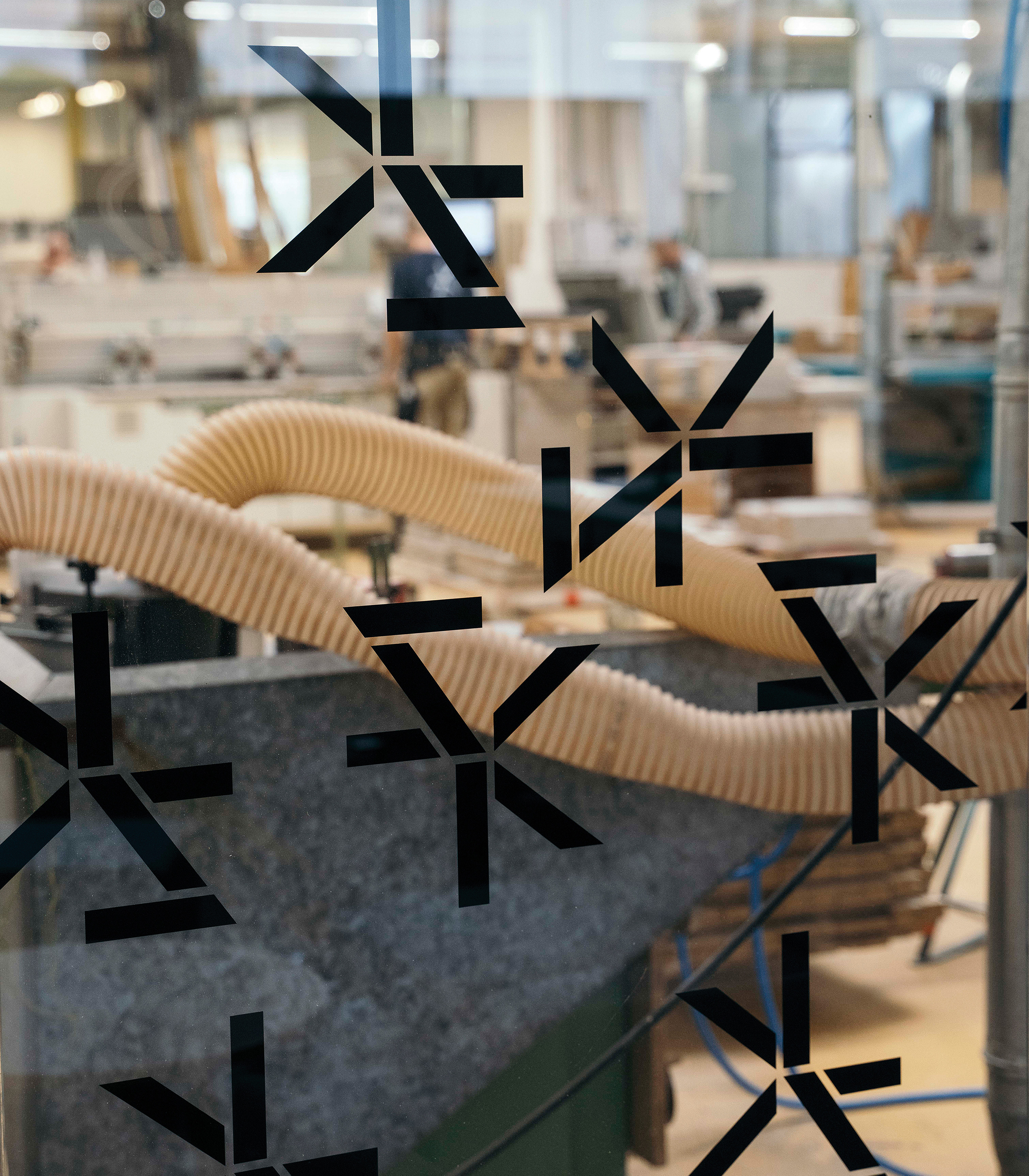 Eine Scheibe mit schwarzen Mustern ist im Vordergrund, wodurch der Blick auf eine Holzwerkstatt im Hintergrund teilweise verdeckt wird. Sichtbar sind Maschinen und Werkstücke, die auf die Bearbeitung warten.