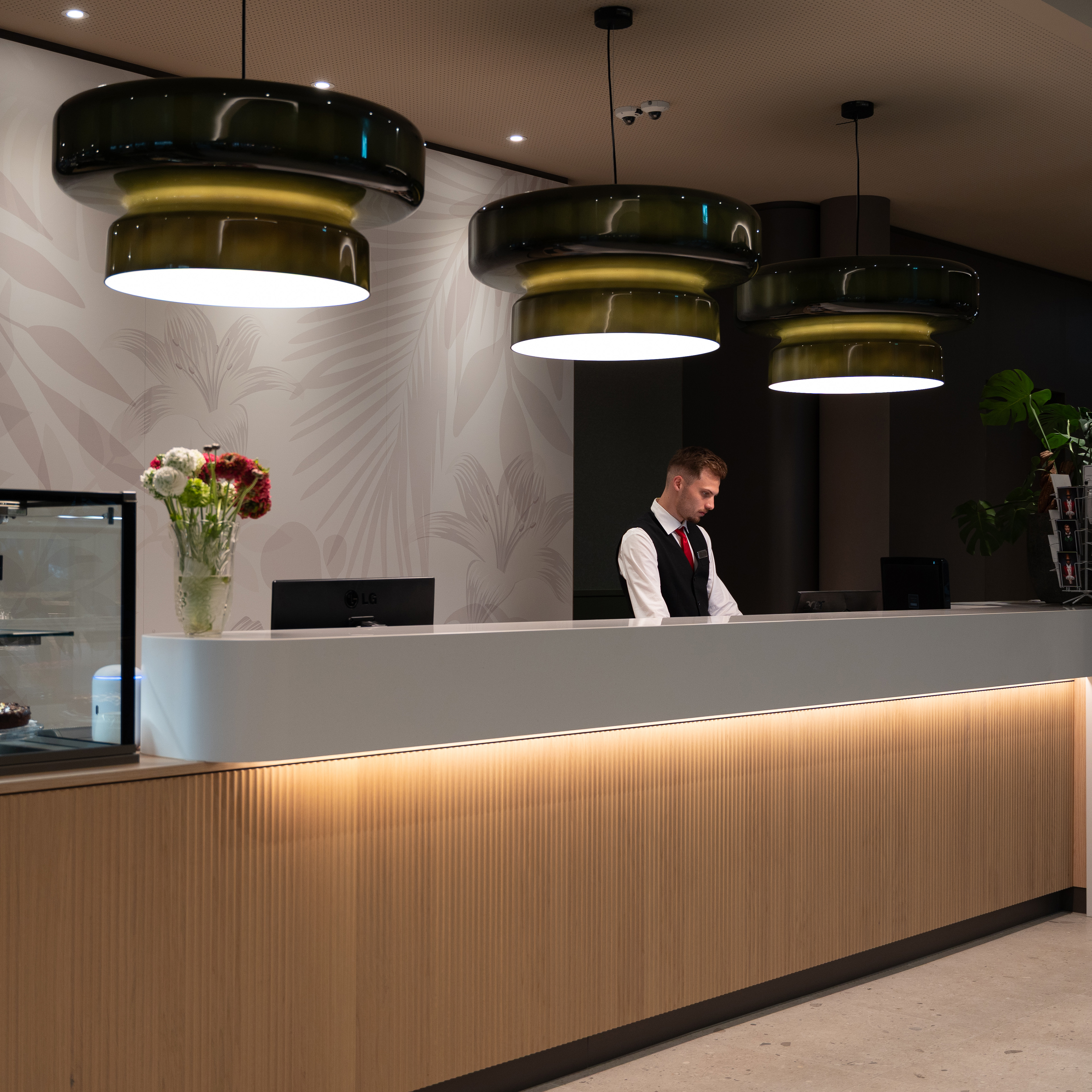 Hotel reception with a modern design, featuring a receptionist, large green pendant lights, floral wallpaper, and a vase of flowers on the counter.