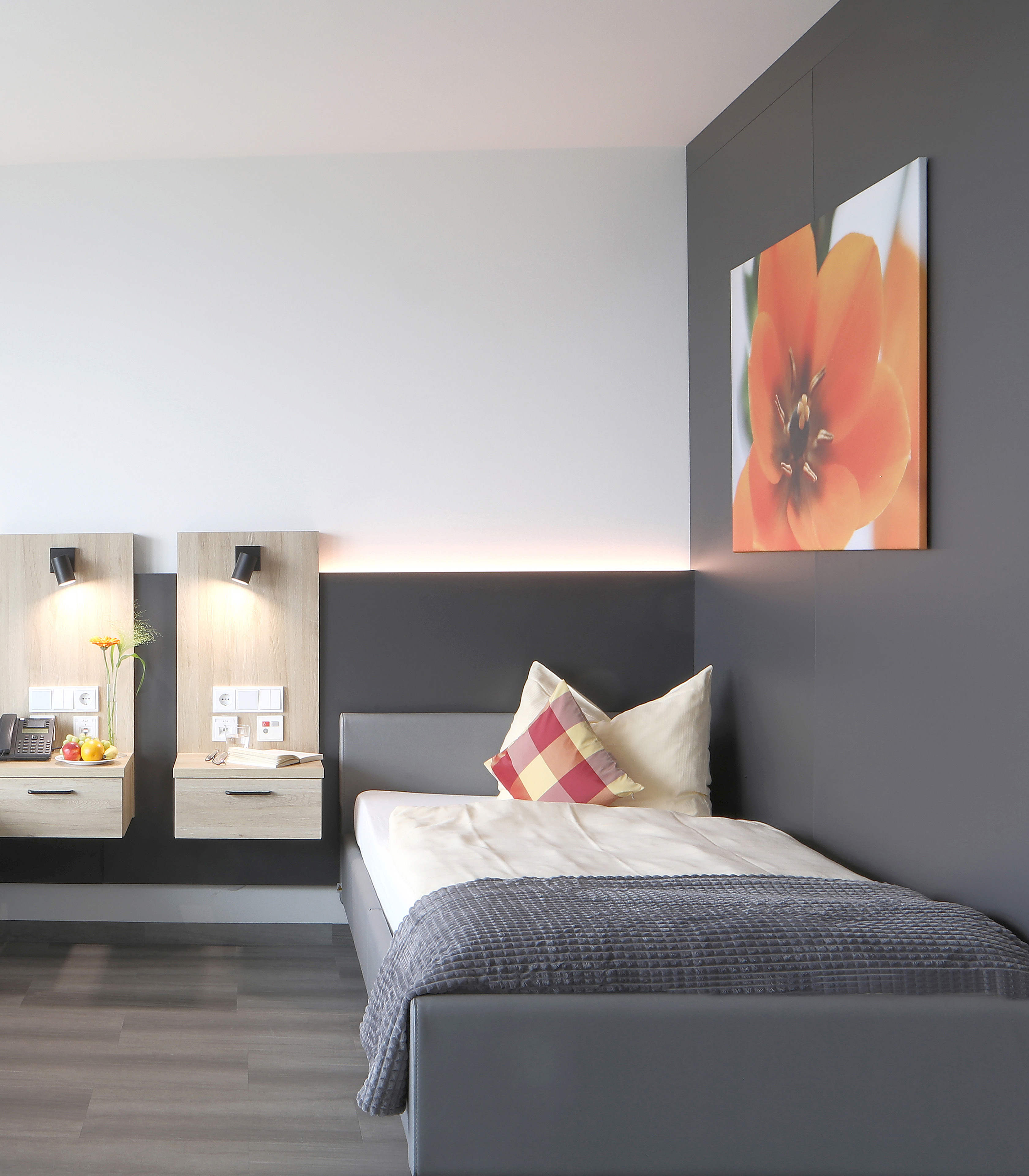Das Bild zeigt ein modernes Schlafzimmer mit einem Einzelbett, einem Kissen in Rot- und Cremetönen sowie einem dunkelgrauen Wandhintergrund. An der Wand hängt ein Bild einer orangefarbenen Blume, und es gibt zwei Nachtkästchen mit Lampen und einer kleinen Vase mit einer Blume.